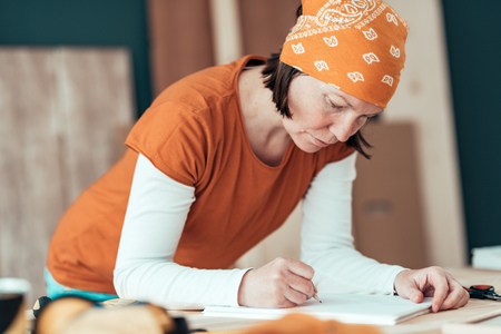 Female carpenter wearing bandanna is sketching project draft on clipboard note paper in her small business woodwork workshopの写真素材
