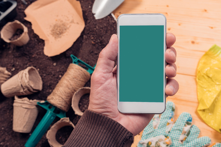 Smartphone screen mock up for gardening and seeding app, mobile phone in gardener's hand over tools and equipment on potting soilの写真素材