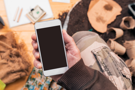 Smartphone mock up in hands of organic gardener, farmer using mobile phone with blank screen over gardening and farming equipmentの写真素材