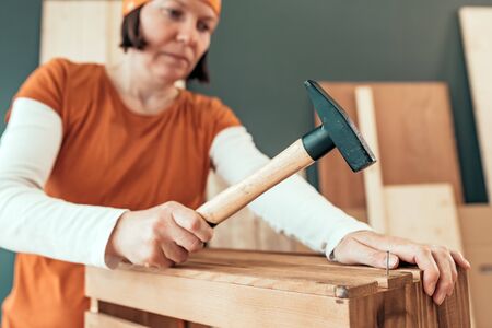 Female carpenter hammering nail into wooden crate in small business woodwork workshop, selective focusの写真素材