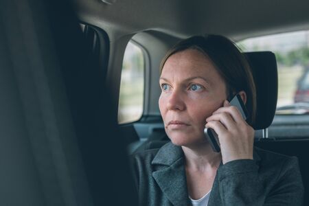 Doubtful businesswoman talking on mobile phone on car back seat, business on the move concept....
