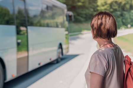Female hiker looking at bus passing by her on road through countryside landscape on sunny summer dayの写真素材