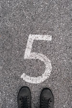 Man standing above number five imprint on asphalt road. Male feet in casual sneakers from above.
