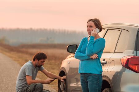 Couple repairing car flat tire on the road in autumn sunset and calling for roadside assistance,...