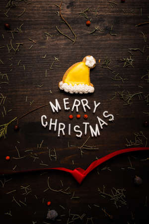 Merry Christmas text with gingerbread Santa Claus hat, flat lay top view on dark wooden backgroundの写真素材