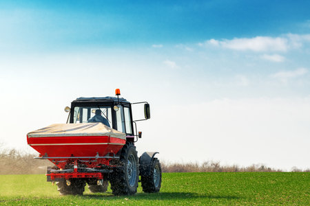 Unrecognizable farmer in agricultural tractor is fertilizing wheat crop field with NPK fertilizer nutrientsの写真素材