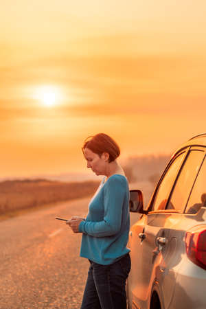 Woman text messaging roadside assistance help after car broke down on countryside road in autumn sunset, selective focus