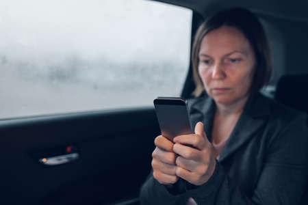 Businesswoman typing text message on mobile phone while sitting on car backseat and commuting to work, selective focusの写真素材