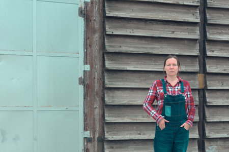 Portrait of female farmer in front of the farmhouse shed. Woman dressed in plid shirt and denim...