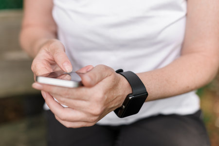 Female hiker using smartphone and smart watch while resting on bench in park, selective focusの写真素材