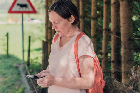 Female hiker text messaging on mobile phone by the road through countryside on sunny summer day...