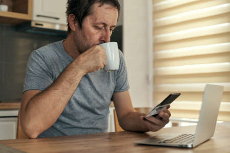 Man shopping online from home with mobile phone and credit card, adult male drinking coffee and...