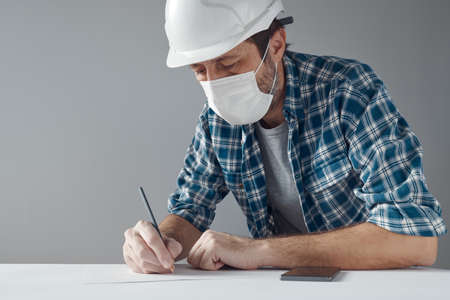 Project engineer with protective face mask writing notes on paper - performing calculations and...
