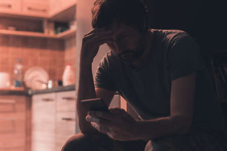 Depressed man typing text message on mobile phone in dark room, selective focusの写真素材