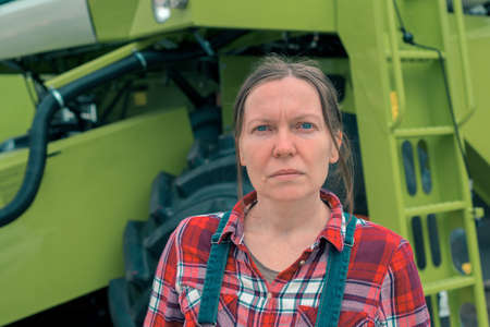 Female farmer posing in front of combine harvester. Serious woman farm worker with agricultural...
