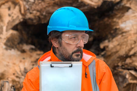 Forestry technician posing with clipboard notepad next to a tree log in forest, confident male...