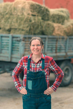 Female farmer posing in front of hay wagon. Portrait of woman farm worker in plaid shirt and bib overalls by the tractor trailer filled with dairy farm livestock feed hay bales.の写真素材