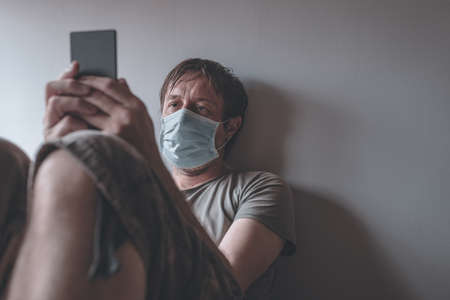 Man in self-isolation during virus outbreak using mobile phone. Worried male person with protective surgical mask, holding smartphone in stay at home concept, selective focusの写真素材