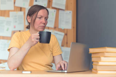Female freelancer working on laptop computer and drinking coffee in home office, selective focus