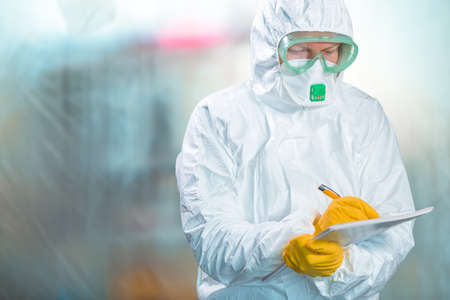 Female epidemiologist in protective clothing writing research report in virus quarantine, medical...