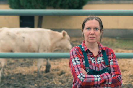 Proud successful farmer at cow dairy farm. Portrait of adult caucasian female farm worker wearing plaid shirt and jeans bib overalls next to the herd in cow shed.の写真素材