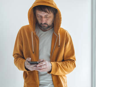 Man with yellow zip hooded sweatshirt using smartphone standing by the window as copy space