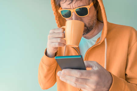 Man drinking coffee and using mobile phone in morning, adult male with yellow zip hoodie holding coffee cup and smartphone, selective focusの写真素材