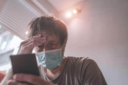 Worried man using mobile phone in home quarantine self-isolation for having Covid-19 coronavirus symptoms, selective focusの写真素材