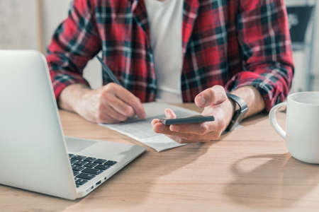 Freelancer writing notes and using smartphone in home office interior, selective focus