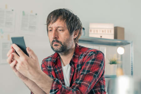 Concerned freelancer reading text message on mobile phone in his small business home office,...