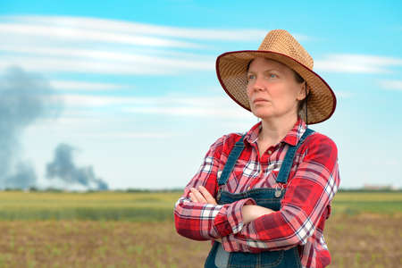 Concerned female farmer in corn field looking at black smoke on horizon, concept of insurance in agriculture and farmingの写真素材
