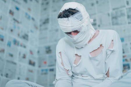 Victim of tabloid journalism and fake news infodemic, conceptual image of man wrapped in bandages surrounded by walls covered with daily newspaper pagesの写真素材
