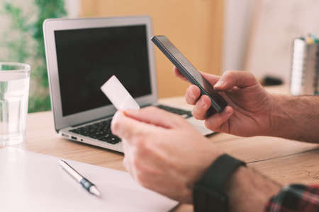 Online shopping from home office using smartphone and credit card, close up of male hands in...