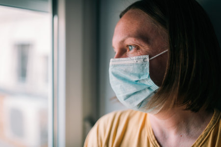 Woman in self-isolation during virus outbreak looking through window. Worried female person with protective surgical mask, selective focusの写真素材