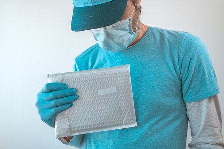 Delivery man posing with parcel package in protective clothing wearing baseball cap, protective mask...