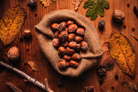 Top view of shelled hazelnuts in environmentally eco friendly burlap sack on rustic wooden table decorated with dry autumn leaves and branchesの写真素材