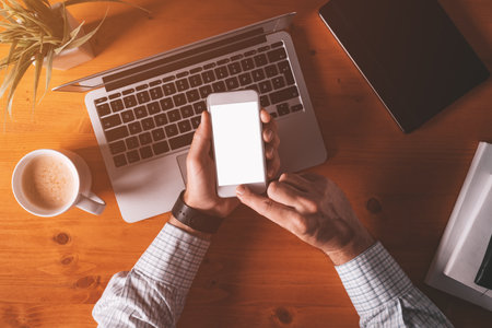 Smartphone mock up overhead shot, businessman holding mobile phone with blank screen over office desk and laptop computerの写真素材