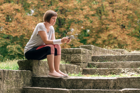 Female jogger texting on smartphone after running in park while sitting on concrete stairsの写真素材