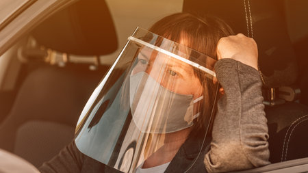 Female taxi driver with protective face mask and plastic visor waiting in car