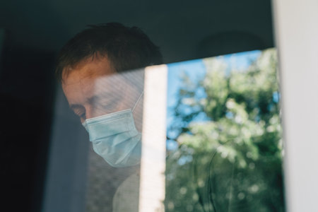 Worried man with protective face mask in self-isolation home quarantine during coronavirus outbreak, portrait of adult male person behind windowの写真素材