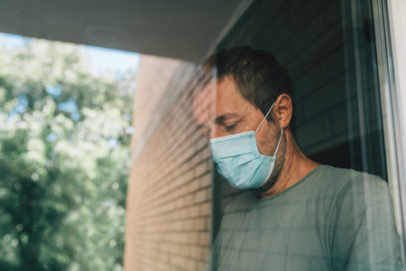 Worried man with protective face mask in self-isolation home quarantine during coronavirus outbreak, portrait of adult male person behind windowの写真素材