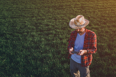 Wheat farmer using drone remote controller in wheatgrass field. Aerial view of male farm worker observing cultivated farmland from drone pov.の写真素材