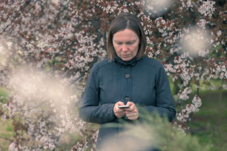 Woman text messaging on mobile phone under the blossoming cherry tree in springtime