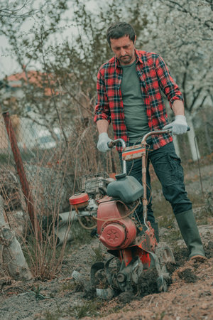 Farmer performing garden tillage with an old motor cultivator, preparing soil for homegrown produce growing, selective focusの写真素材