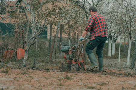 Farmer performing garden tillage with an old motor cultivator, preparing soil for homegrown produce growing, selective focusの写真素材
