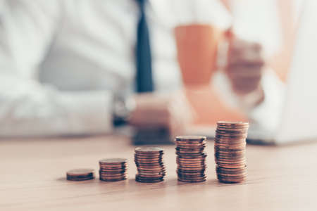 Satisfied businessman drinking coffee with coin stack in front, close up with selective focusの写真素材
