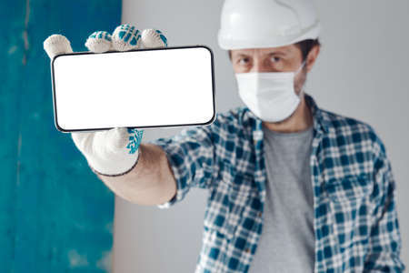 Construction engineer with protective face mask holding smartphone with blank mock up screen for...
