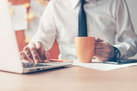 Businessman drinking coffee and using laptop computer in the office, close up with selective focus