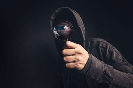 Bizarre hooded spooky person with magnifying glass, focus on enlarged eye peeking at youの写真素材