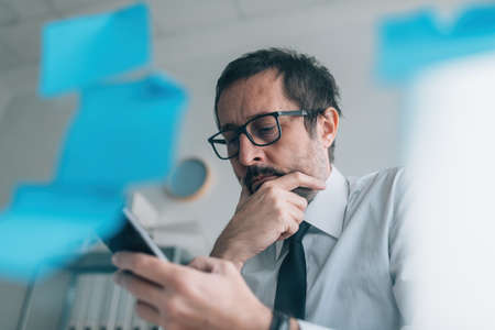 Considerate businessman thinking of text message reply and looking at mobile phone in office, selective focusの写真素材